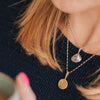 A 9ct gold full moon-shaped pendant necklace worn by a blonde lady in a navy jumper, stacked with a chunky shell necklace
