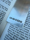 A close-up of a brushed silver metal bookmark resting on open book pages. The bookmark features engraved hallmarks including 'LK', '925', a lion passant, and 'B'.
