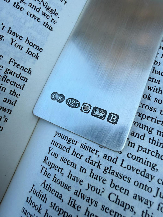 A close-up of a brushed silver metal bookmark resting on open book pages. The bookmark features engraved hallmarks including 'LK', '925', a lion passant, and 'B'.