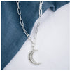 A silver crescent moon pendant hanging from a trace chain necklace, displayed against a denim background.