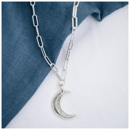 A silver crescent moon pendant hanging from a trace chain necklace, displayed against a denim background.