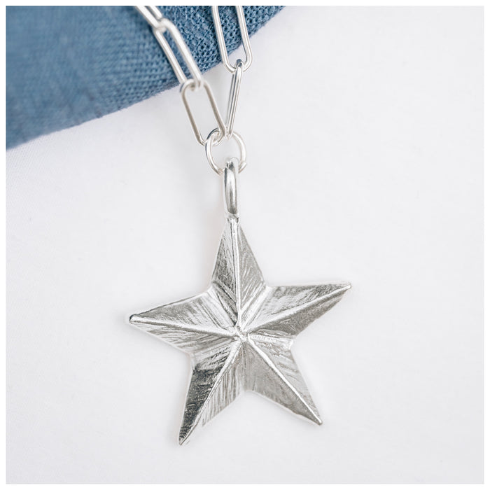 A silver Guiding Star pendant hanging from a trace chain necklace, displayed against a denim background.