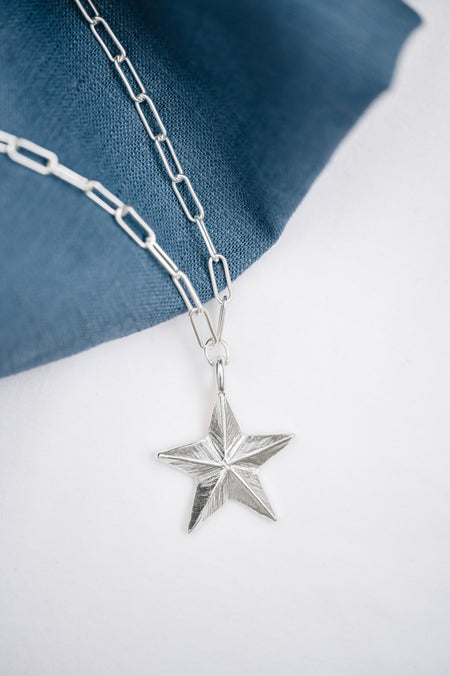 A silver necklace featuring a guiding star pendant, laid out on a blue fabric surface.