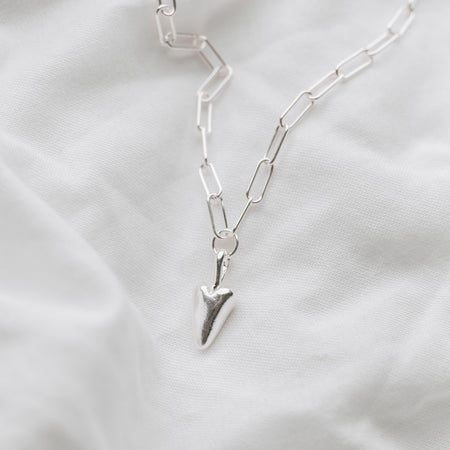 A silver trace chain necklace with a heart-shaped pendant, displayed on a textured white background.