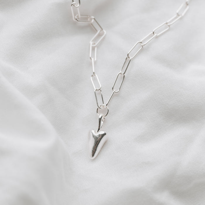 A silver trace chain necklace with a heart-shaped pendant, displayed on a textured white background.