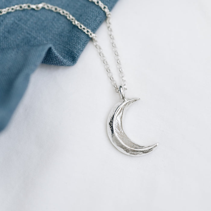 A crescent moon pendant made from recycled sterling silver, hanging from a silver belcher chain on a white background, with a blue denim fabric beside it.