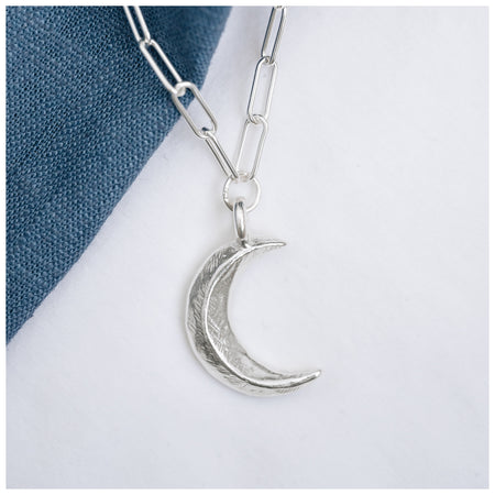 A silver crescent moon pendant hanging from a trace chain necklace, displayed against a denim background.