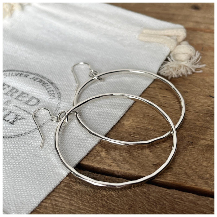 A pair of sterling silver hoop earrings with a hammered effect, displayed on a textured wooden surface.