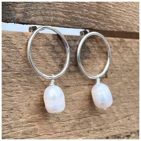 A pair of silver hoop earrings with freshwater pearl drops, displayed against a wooden background.