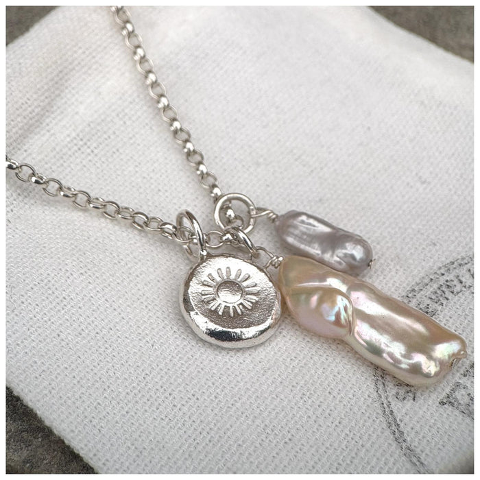 A silver belcher chain necklace with three charms: a small grey pearl, a white/silver stick pearl & a silver pebble with a stamped sunshine design, displayed against a wooden background.