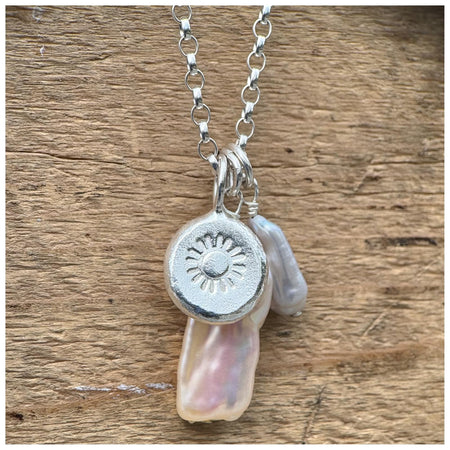 A silver belcher chain necklace with three charms: a small grey pearl, a white/silver stick pearl & a silver pebble with a stamped sunshine design, displayed against a wooden background.