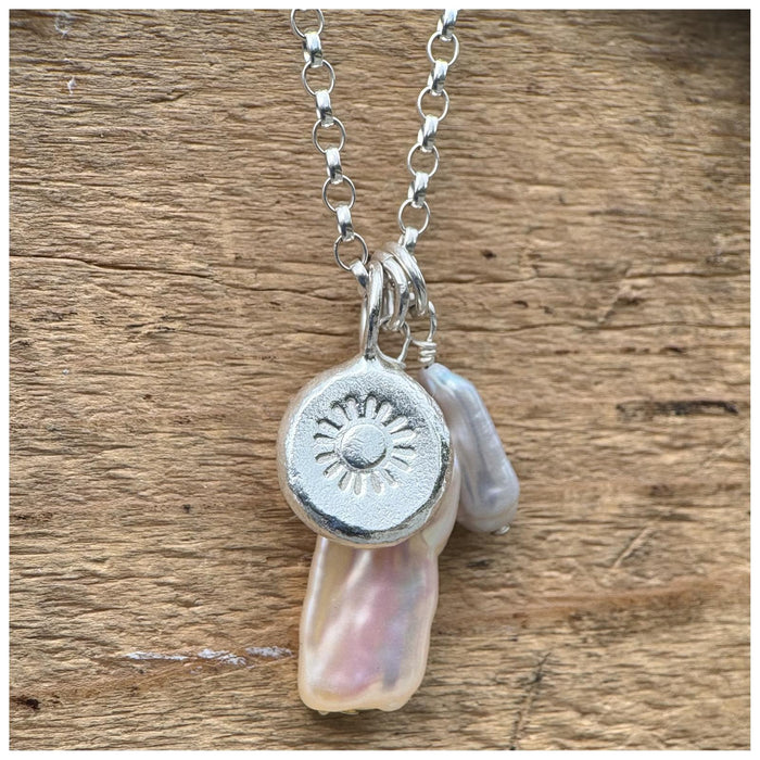 A silver belcher chain necklace with three charms: a small grey pearl, a white/silver stick pearl & a silver pebble with a stamped sunshine design, displayed against a wooden background.