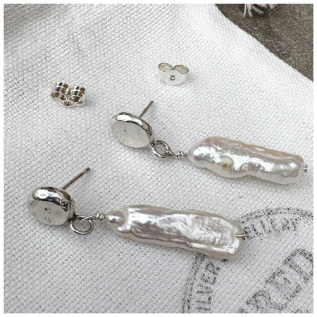 Sterling silver earrings with a silver pebble stud and a delicate stick pearl hanging from it, both laying on a  white linen bag with the Fred & Emily Logo stamped on