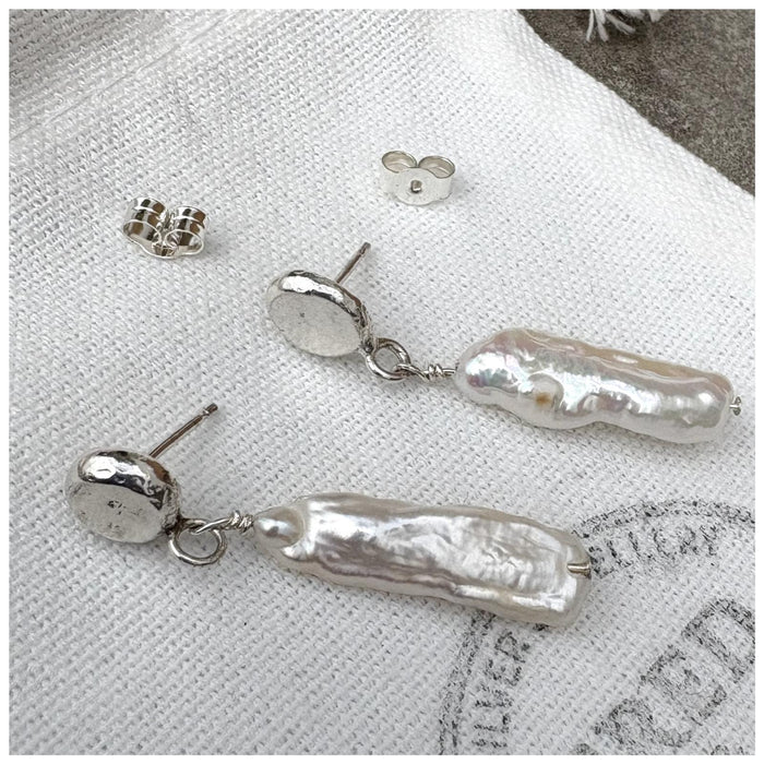 Sterling silver earrings with a silver pebble stud and a delicate stick pearl hanging from it, both laying on a  white linen bag with the Fred & Emily Logo stamped on