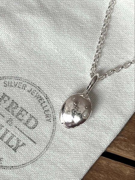 A steriing silver pendant shaped like a pumpkin seed hanging on a chunky chain on a linen bag