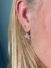 A close-up of a person's ear showcasing silver star-shaped stud earrings and hoop earrings, with blonde hair framing the side of the face against a muted blue-gray background.