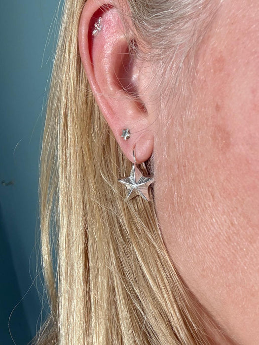 A close-up of a person's ear showcasing silver star-shaped stud earrings and hoop earrings, with blonde hair framing the side of the face against a muted blue-gray background.