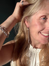 A close-up lifestyle shot of a smiling blonde woman wearing a long, dangling silver earring and a chunky silver chain bracelet, with her hand in her hair against a dark background.