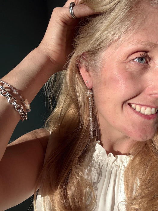 A close-up lifestyle shot of a smiling blonde woman wearing a long, dangling silver earring and a chunky silver chain bracelet, with her hand in her hair against a dark background.
