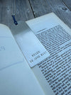 A silver metal bookmark engraved with the name 'HELEN' and the date '28 JUNE 2001', resting on an open book on a weathered wooden surface.