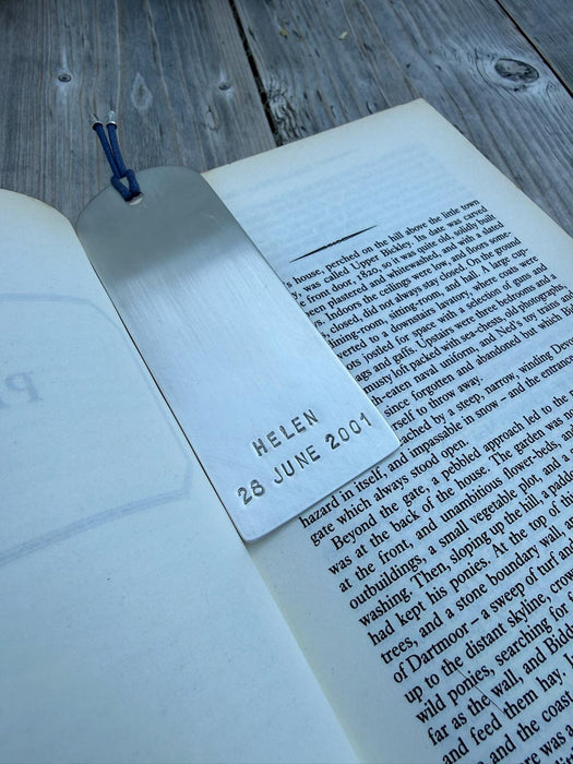 A silver metal bookmark engraved with the name 'HELEN' and the date '28 JUNE 2001', resting on an open book on a weathered wooden surface.