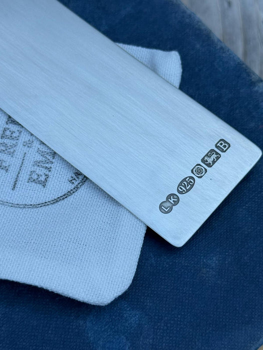 A close-up of a silver metal bookmark with a large hallmark, resting on a white textured cloth over a dark blue fabric background.