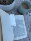 A sterling silver bookmark with a blue string and hallmark, placed in an open book on a light wood surface next to a green ceramic mug and a potted succulent.