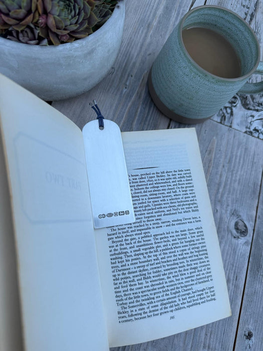 A sterling silver bookmark with a blue string and hallmark, placed in an open book on a light wood surface next to a green ceramic mug and a potted succulent.