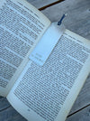 A silver metal bookmark with a blue string, printed with 'HELEN 28 JUNE 2017', resting on the pages of an open book on a weathered wooden surface.