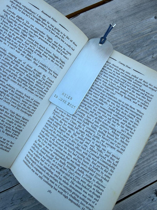 A silver metal bookmark with a blue string, printed with 'HELEN 28 JUNE 2017', resting on the pages of an open book on a weathered wooden surface.