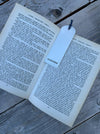 A sterling silver bookmark with a blue ribbon tail rests on an open book spread on a weathered gray wooden surface.