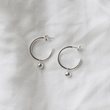 Silver hooped earrings with a small globe of silver hanging from each, laying on a flat white linen background