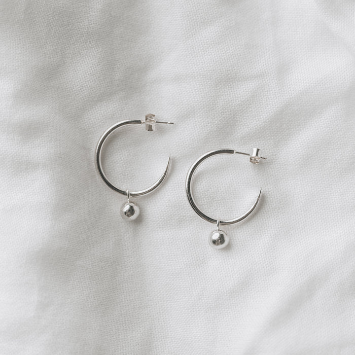 Silver hooped earrings with a small globe of silver hanging from each, laying on a flat white linen background