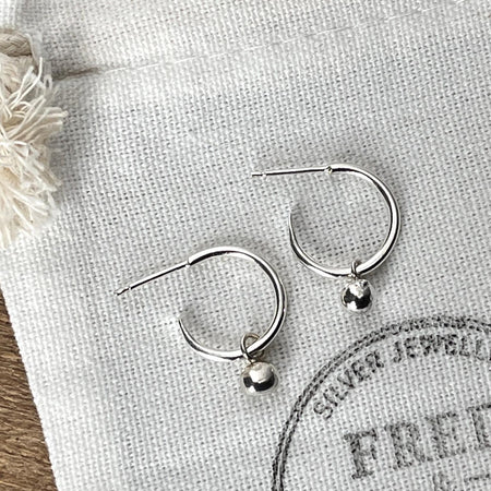 Delicate silver hoops with a small globe of silver hanging from each, displayed on a white linen bag with the Fred & Emily Logo printed on.