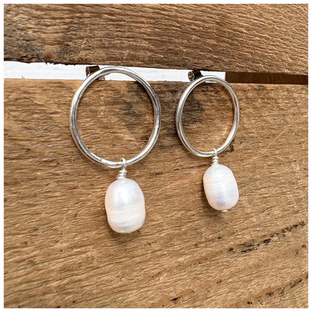 A pair of silver hoop earrings with freshwater pearl drops, displayed against a wooden background.