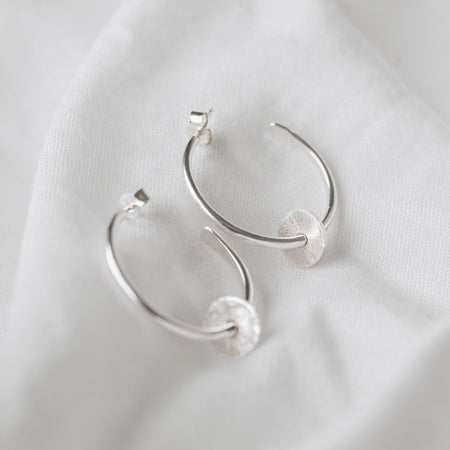 Sterling silver hoops with a textured disc on each, laying on a white linen background with butterfly backs
