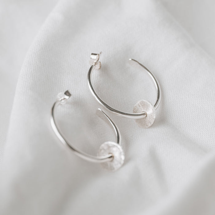 Sterling silver hoops with a textured disc on each, laying on a white linen background with butterfly backs