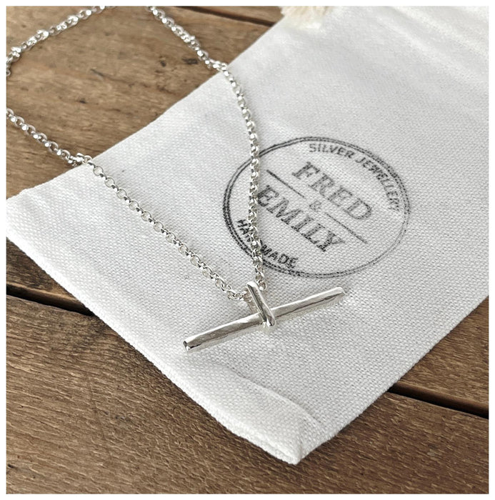 A silver T bar necklace with a hammered finish, laid on a belcher chain on a white linen bag..
