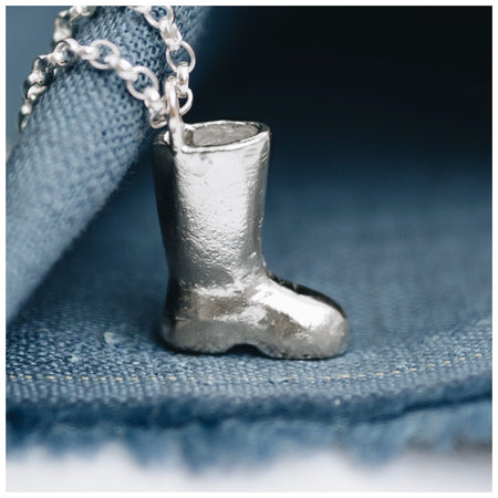A silver wellington boot pendant on a chunky belcher chain, placed on a blue fabric surface.