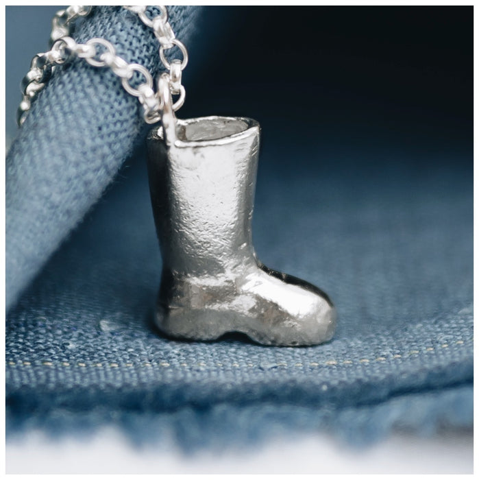 A silver wellington boot pendant on a chunky belcher chain, placed on a blue fabric surface.