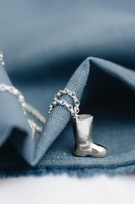 A silver wellington boot pendant on a chunky belcher chain, placed on a blue fabric surface.