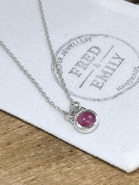 A sterling silver pendant with an oval pink tourmaline stone on an 18'' belcher chain, displayed on a white fabric with the brand mark 'FRED & EMILY'.