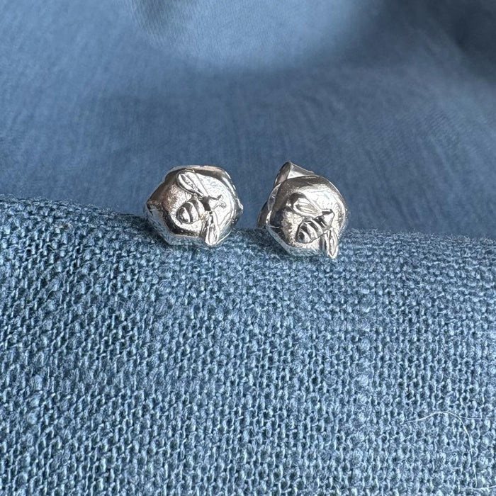 Sterling silver stud earrings with a bee on a piece of silver honeycomb sitting on a blue textured fabric background.