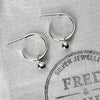 A pair of sterling silver hoop earrings with silver globes attached, displayed on a white fabric with the brand name 'FRED & EMILY' printed on it.