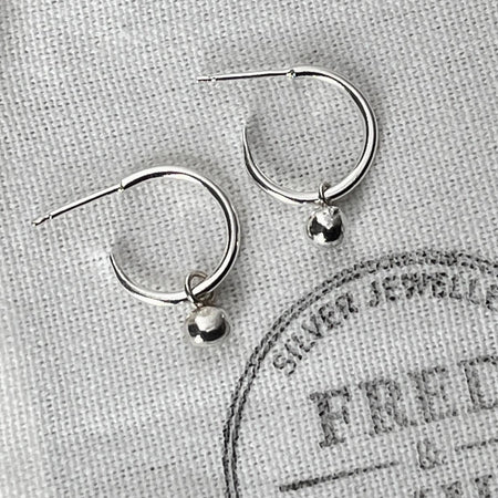 A pair of sterling silver hoop earrings with silver globes attached, displayed on a white fabric with the brand name 'FRED & EMILY' printed on it.