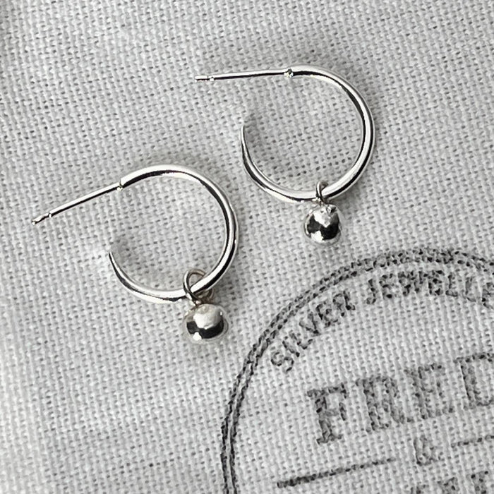 A pair of sterling silver hoop earrings with silver globes attached, displayed on a white fabric with the brand name 'FRED & EMILY' printed on it.