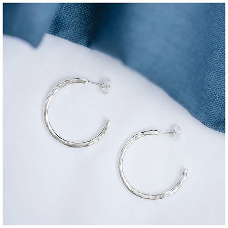 Sterling silver midi hammered hoop with silver butterfly backs sitting on a white background with a blue linen nearby