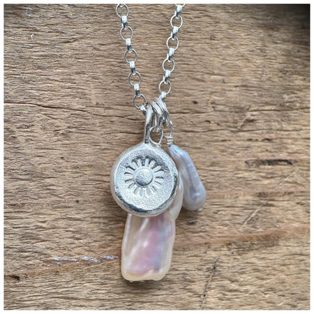 A silver belcher chain necklace with three charms: a small grey pearl, a white/silver stick pearl & a silver pebble with a stamped sunshine design, displayed against a wooden background.