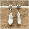 Sterling silver earrings with a silver pebble stud and a delicate stick pearl hanging from it, both on a wooden background