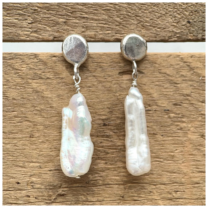 Sterling silver earrings with a silver pebble stud and a delicate stick pearl hanging from it, both on a wooden background